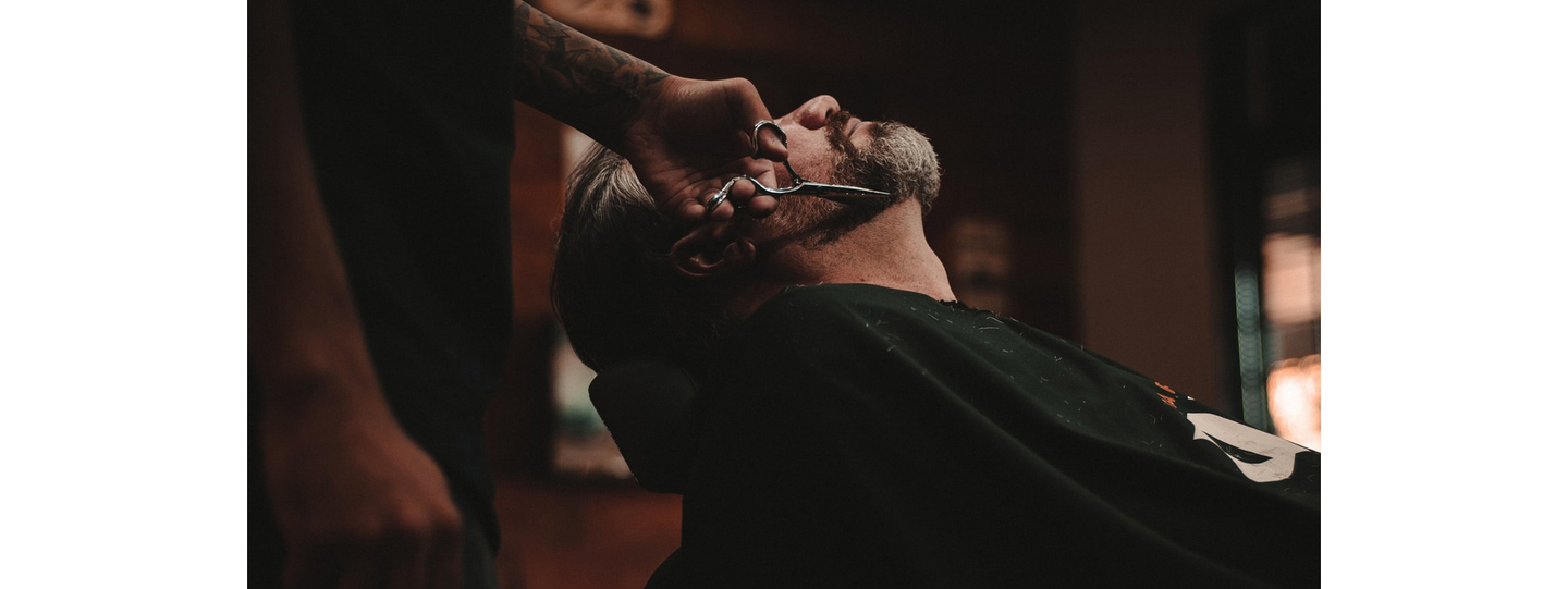 Man getting his beard trimmed with scissors by a barber in a dimly lit setting.